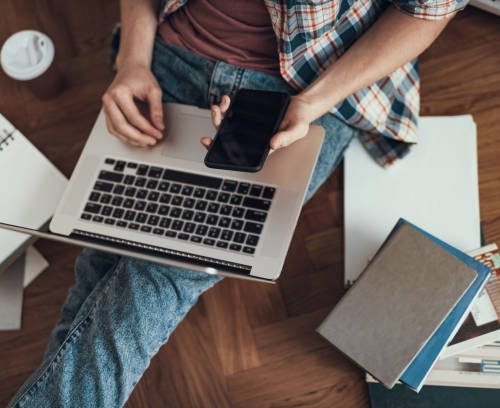 Top view of progressive person holding modern smartphone while sitting on the floor with laptop on the knees